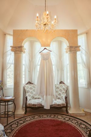 Elegant white wedding dress hanging between classical columns under a crystal chandelier.