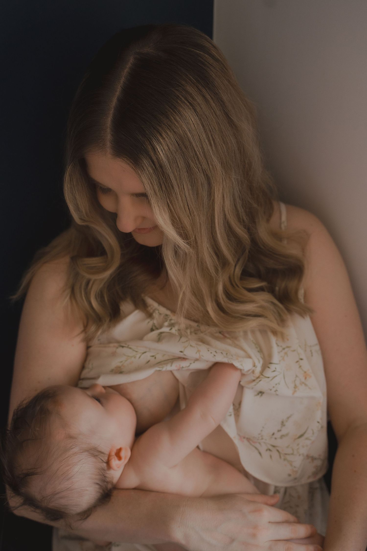 A mother breastfeeding her baby while wrapped together on the bed in soft light, captured during an intimate and gentle lifestyle session at
