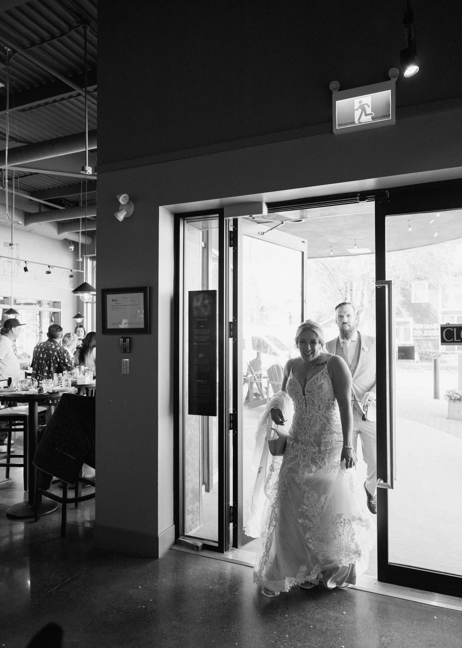 A person in a flowing white wedding dress enters through glass doors of a modern venue in black and white photography.