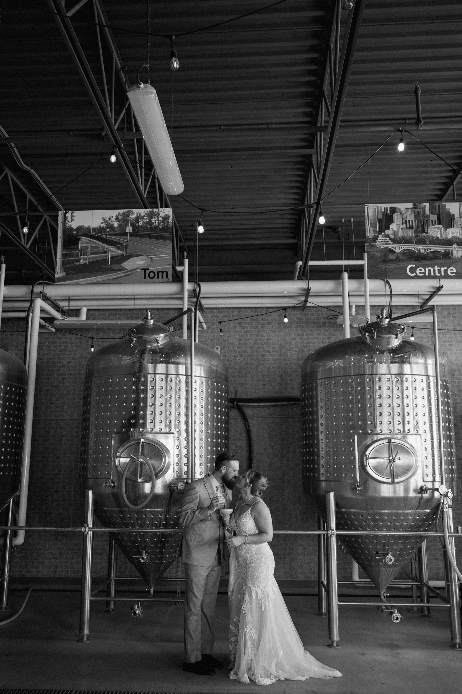 Couple sharing an intimate moment among large steel brewing tanks in an industrial space.