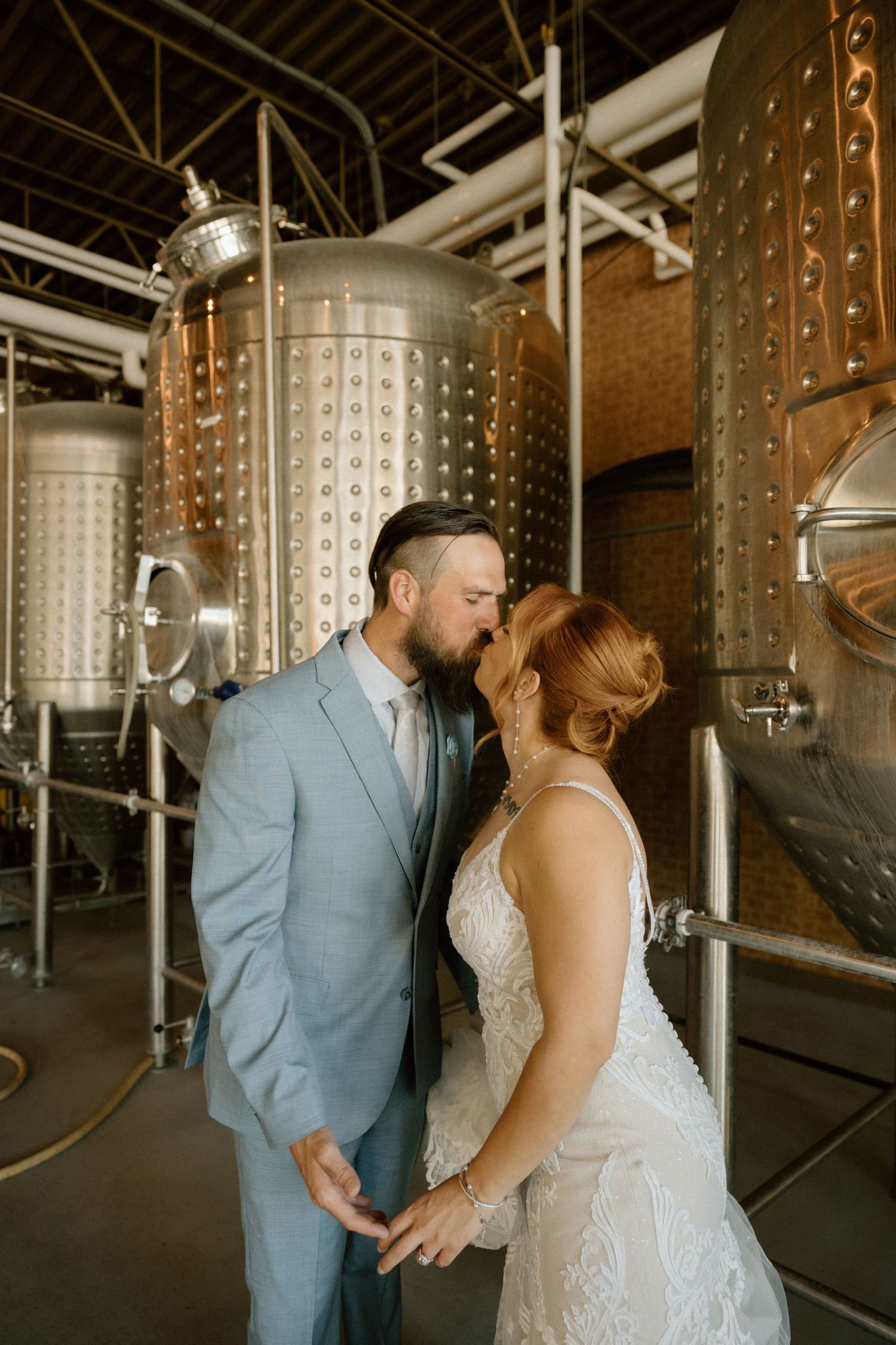 A wedding couple shares an intimate moment in front of industrial steel brewing tanks.