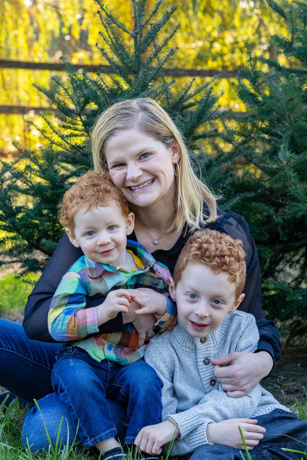 A family sits together on grass near evergreen trees with golden sunlight filtering through the branches in the background.