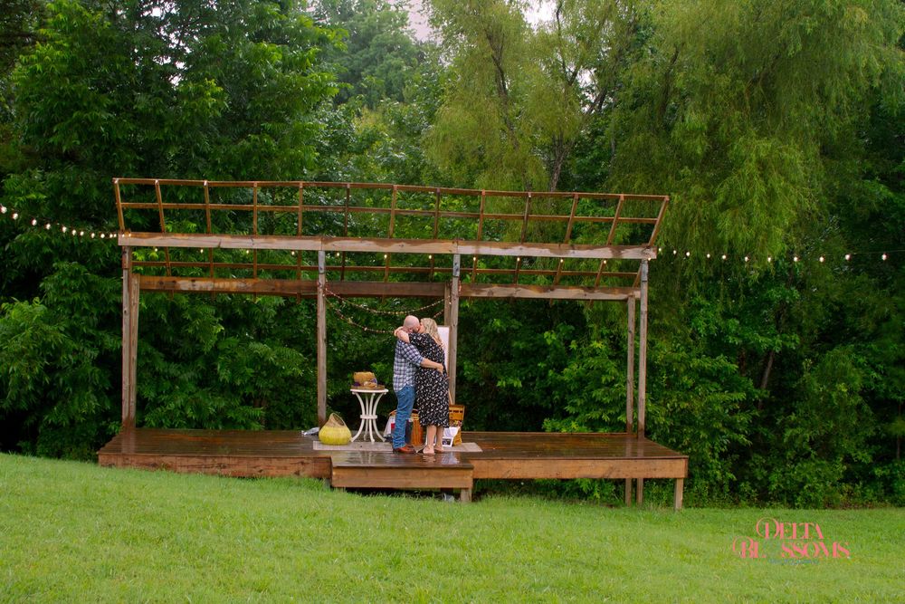 a newly engaged couple standing on a rustic stage with trees in the background
