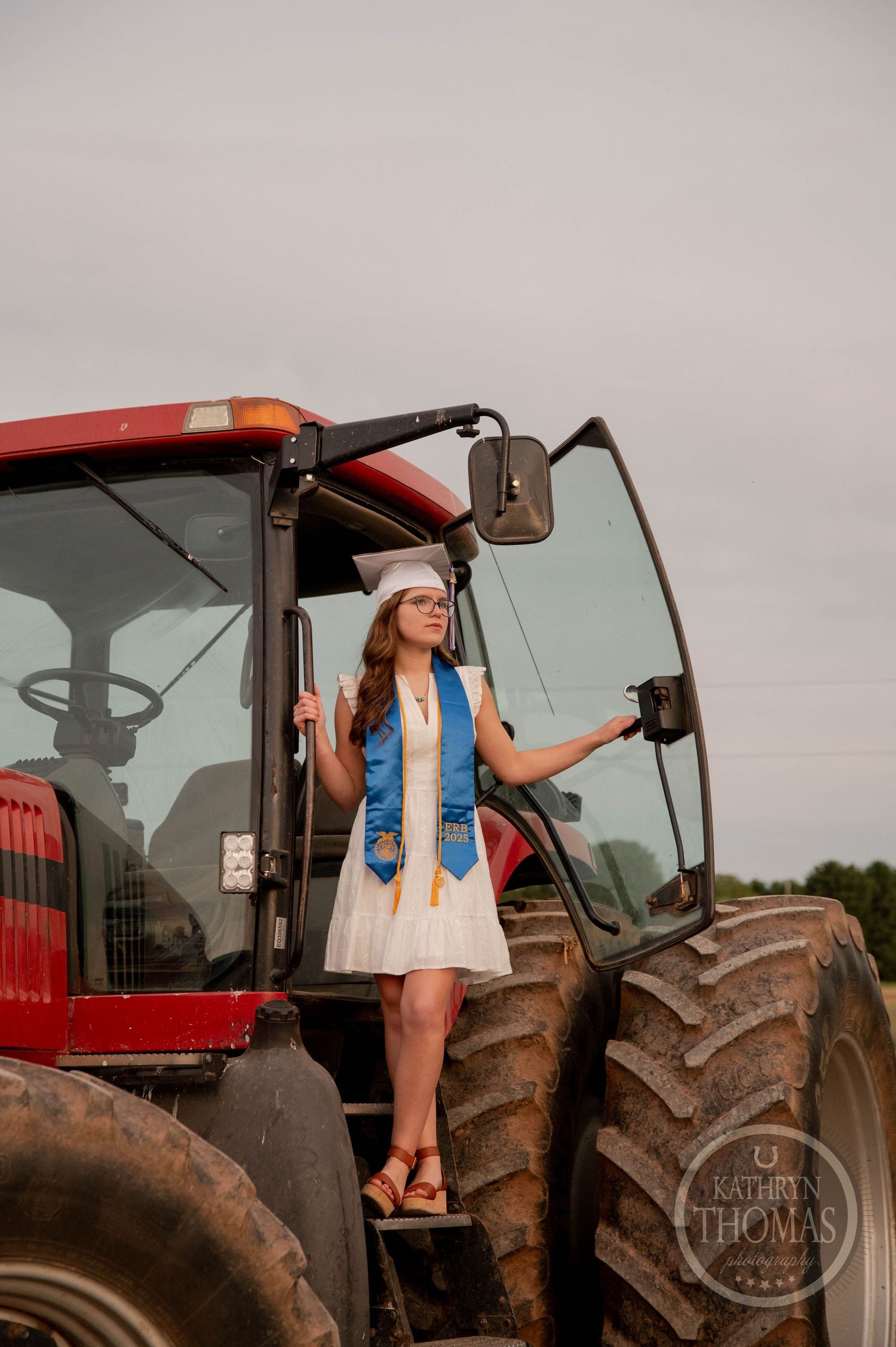 A young woman in a blue vest and white dress stands on the steps of a large red tractor at sunset.