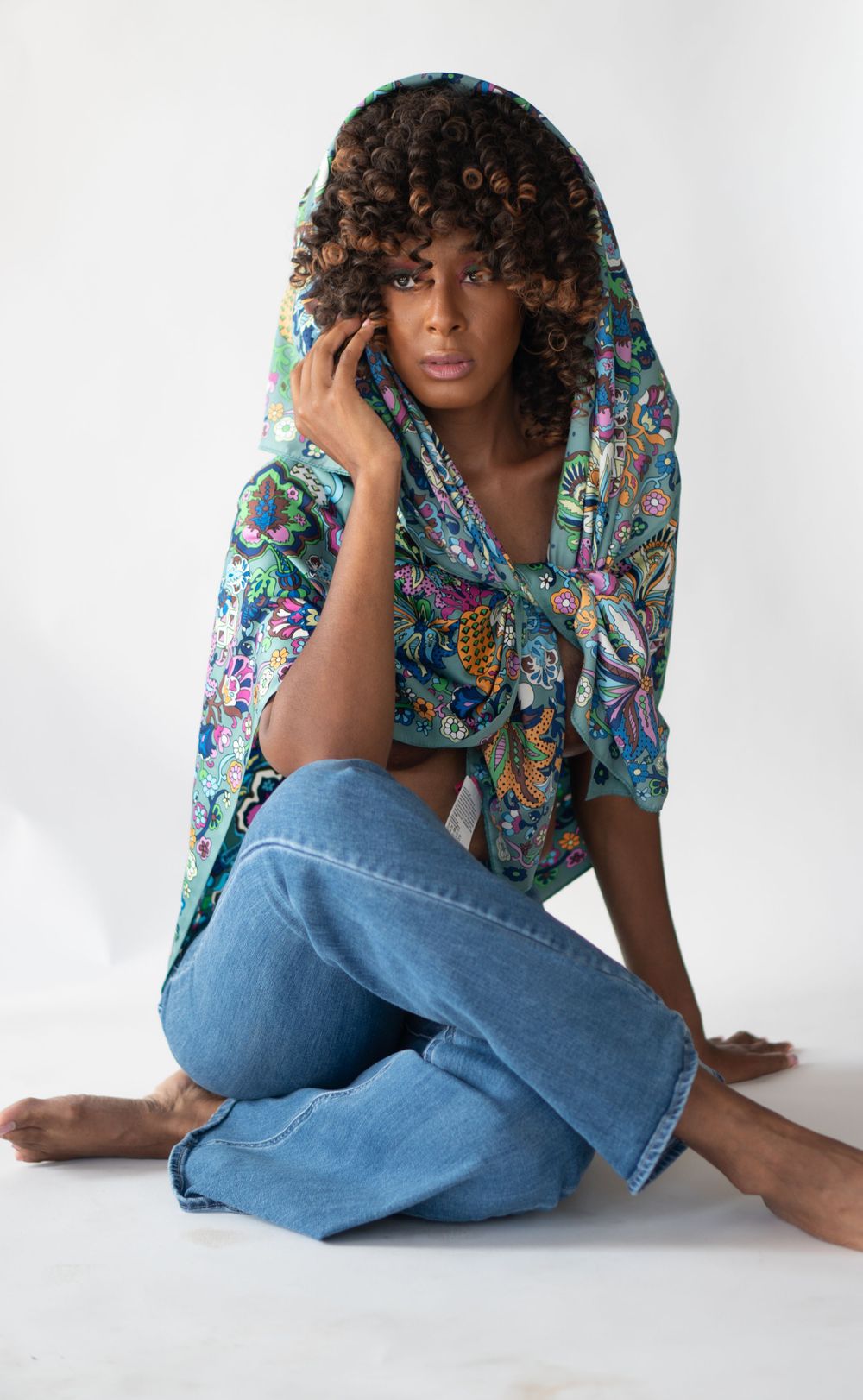 Model in floral print scarf, used as a hoodi,  and jeans poses casually against white background.