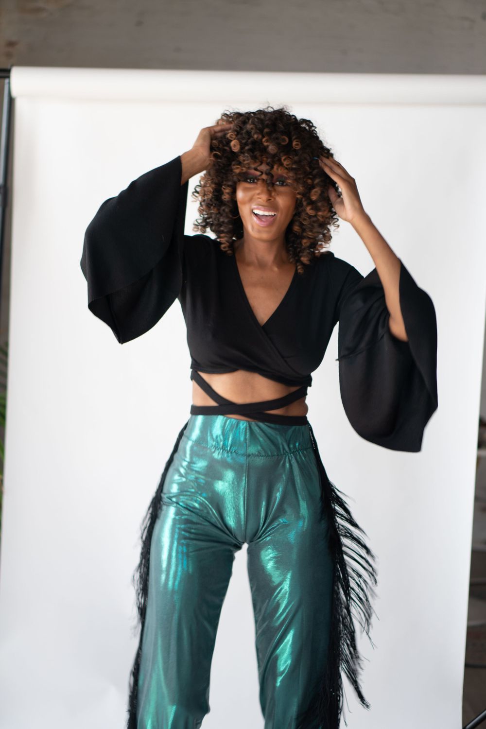 Dynamic fashion pose in black crop top and metallic green pants with dramatic leather cowboy frills.