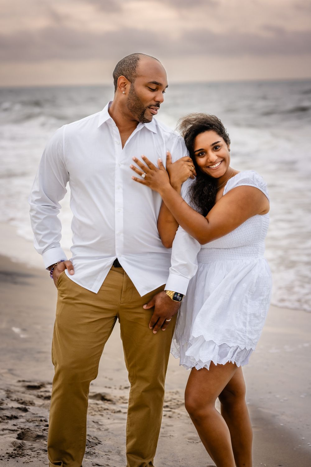 A couple shares a romantic moment on the beach at sunset wearing white and tan attire.