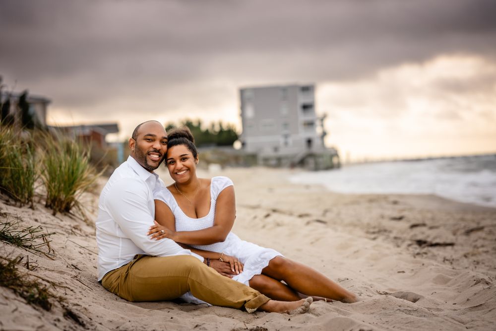 A couple sits together on a sandy beach with an ocean view and cloudy sky in the background.