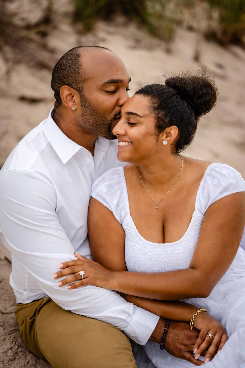 A couple shares an intimate moment on the beach while dressed in white and khaki clothing.