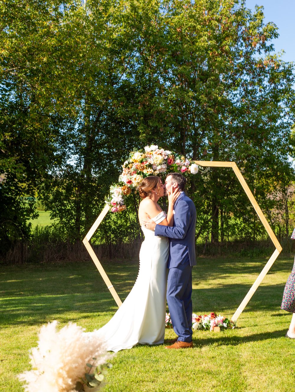 A wedding ceremony takes place under a hexagonal floral arch against a backdrop of lush green trees on a sunny day.