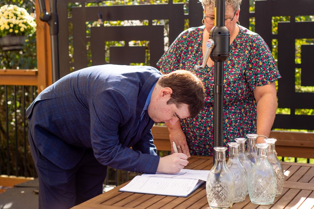 A person in a navy suit signs documents at a wooden table during an outdoor event.