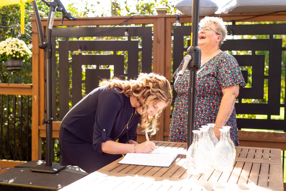 Wedding guests signing documents at an outdoor ceremony with decorative wooden backdrop.