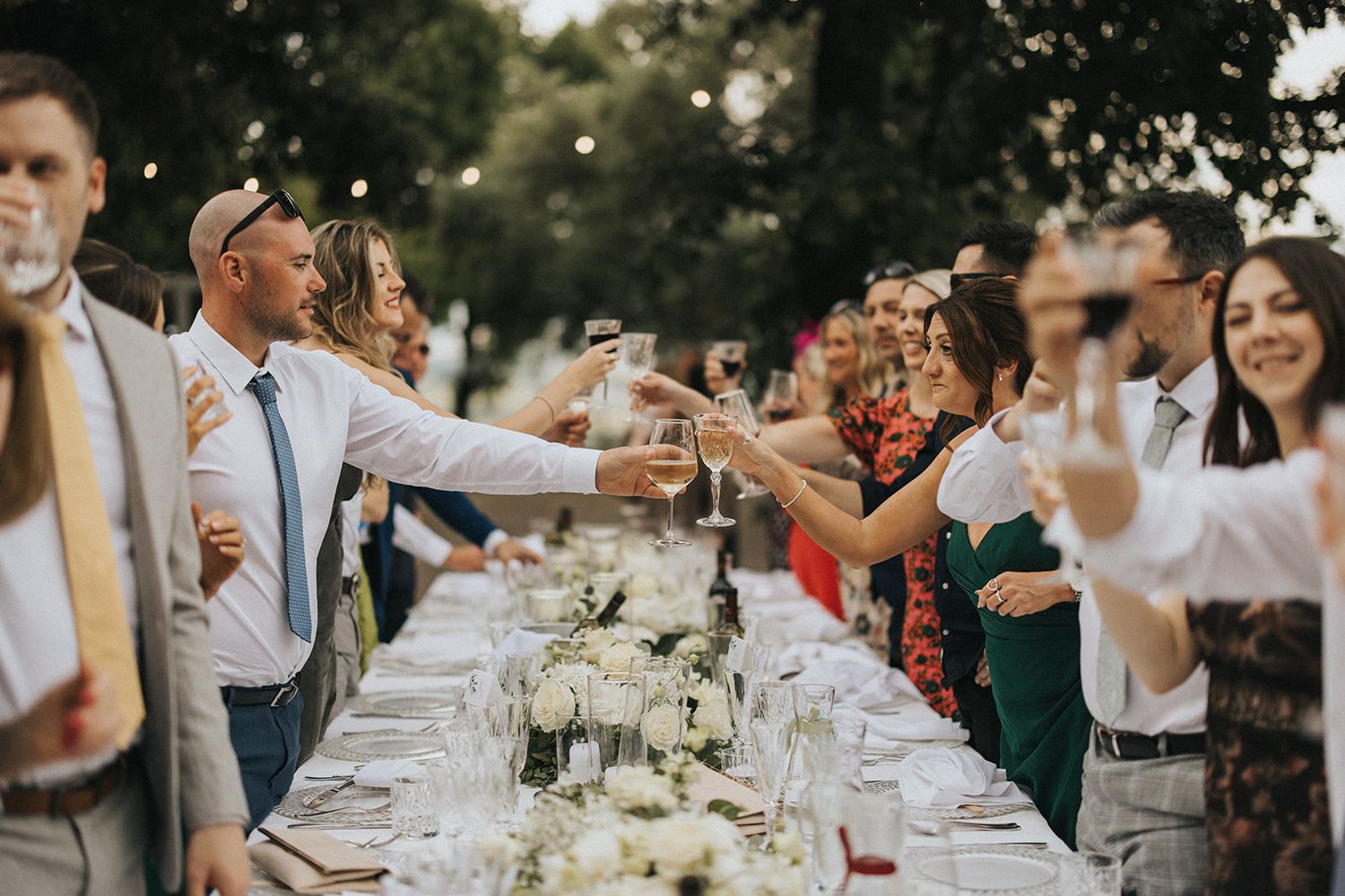 Wedding guests raise their glasses in celebration at a long outdoor dining table.