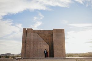 Couple standing in a large geometric concrete sculpture in a desert landscape under a partly cloudy sky.