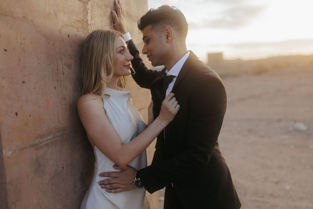 Couple in wedding attire sharing a romantic moment against a rustic wall at sunset in a desert setting.