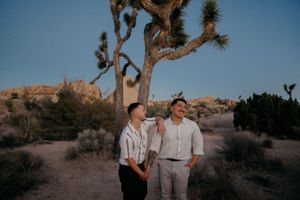 Two people stand together at sunset in Joshua Tree National Park with desert landscape and Joshua trees in background.