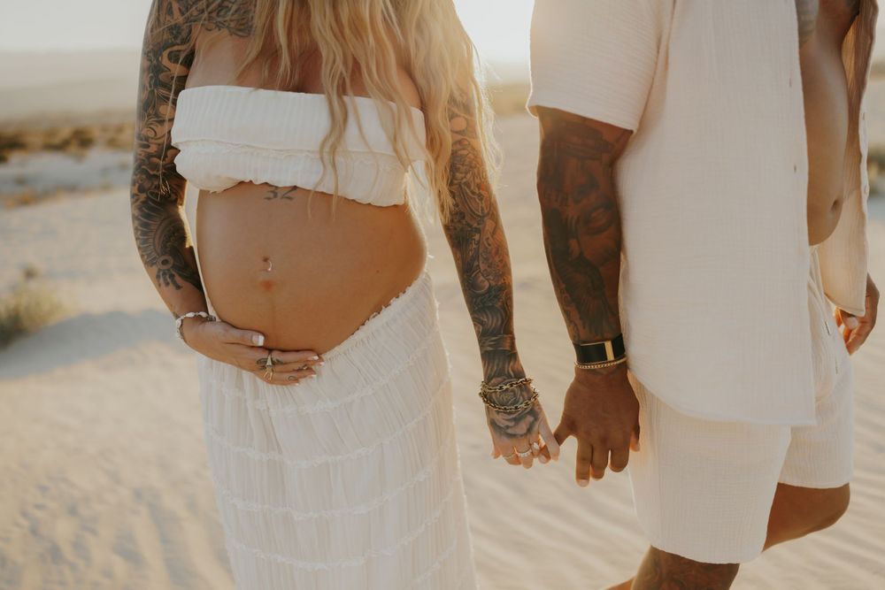 A couple in white clothing holding hands on a sandy beach showing a pregnant belly and tattoo covered arm.
