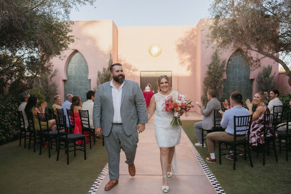 Bride and groom walk down outdoor wedding aisle at pink venue, bride holding colorful bouquet, guests seated on black chairs.