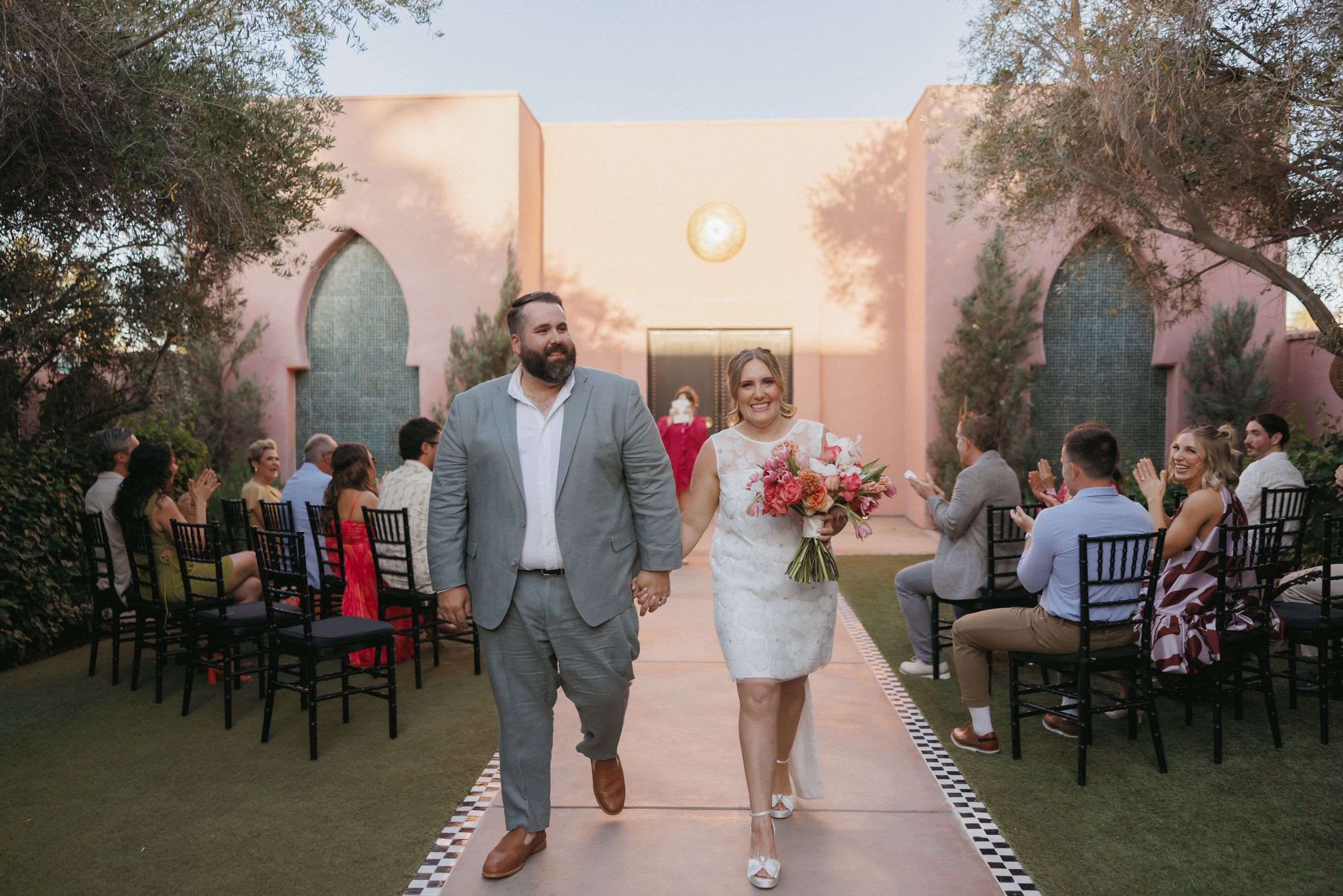 Bride and groom walk down outdoor wedding aisle at pink venue, bride holding colorful bouquet, guests seated on black chairs.