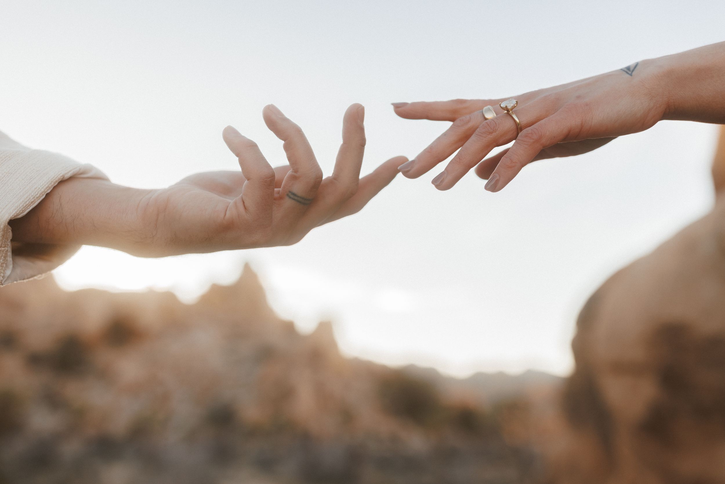 Two hands reaching toward each other against a soft sky, one adorned with a gold ring, evoking connection and romance.