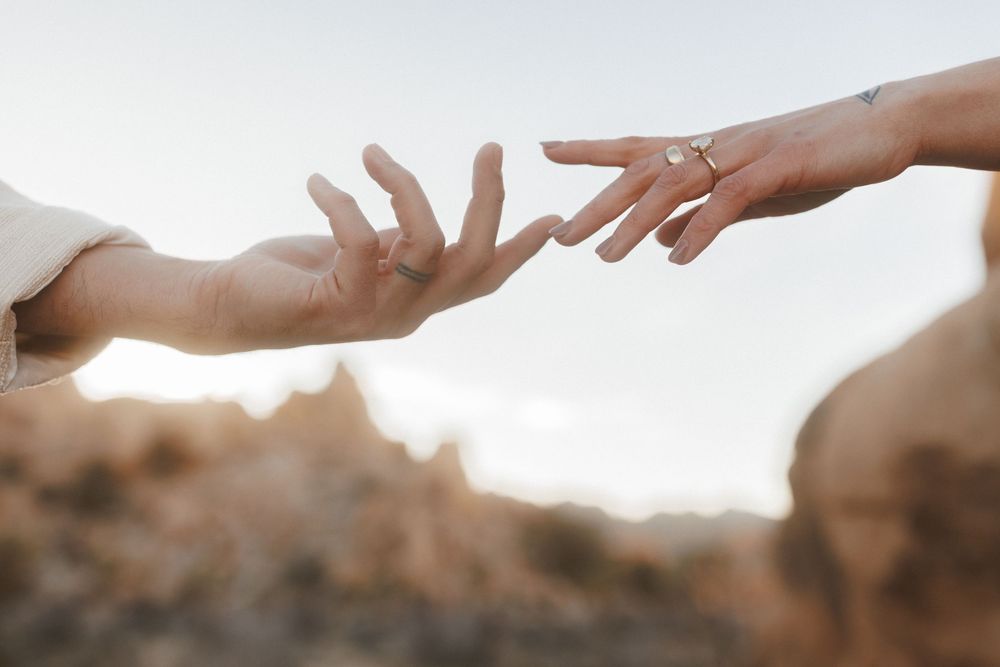 Two hands reaching toward each other against a soft sky, one adorned with a gold ring, evoking connection and romance.