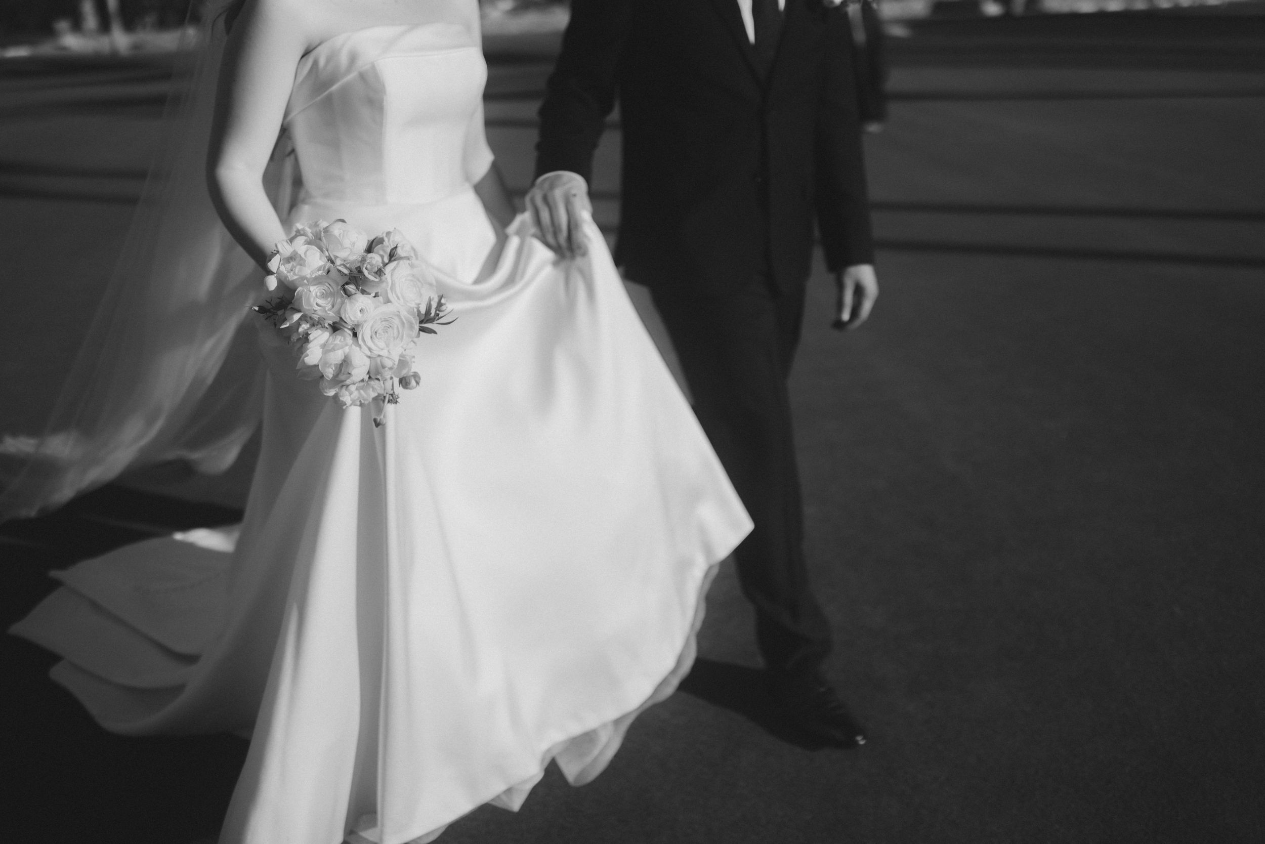 Black and white photo of bride and groom walking together, bride holding bouquet and flowing wedding dress.