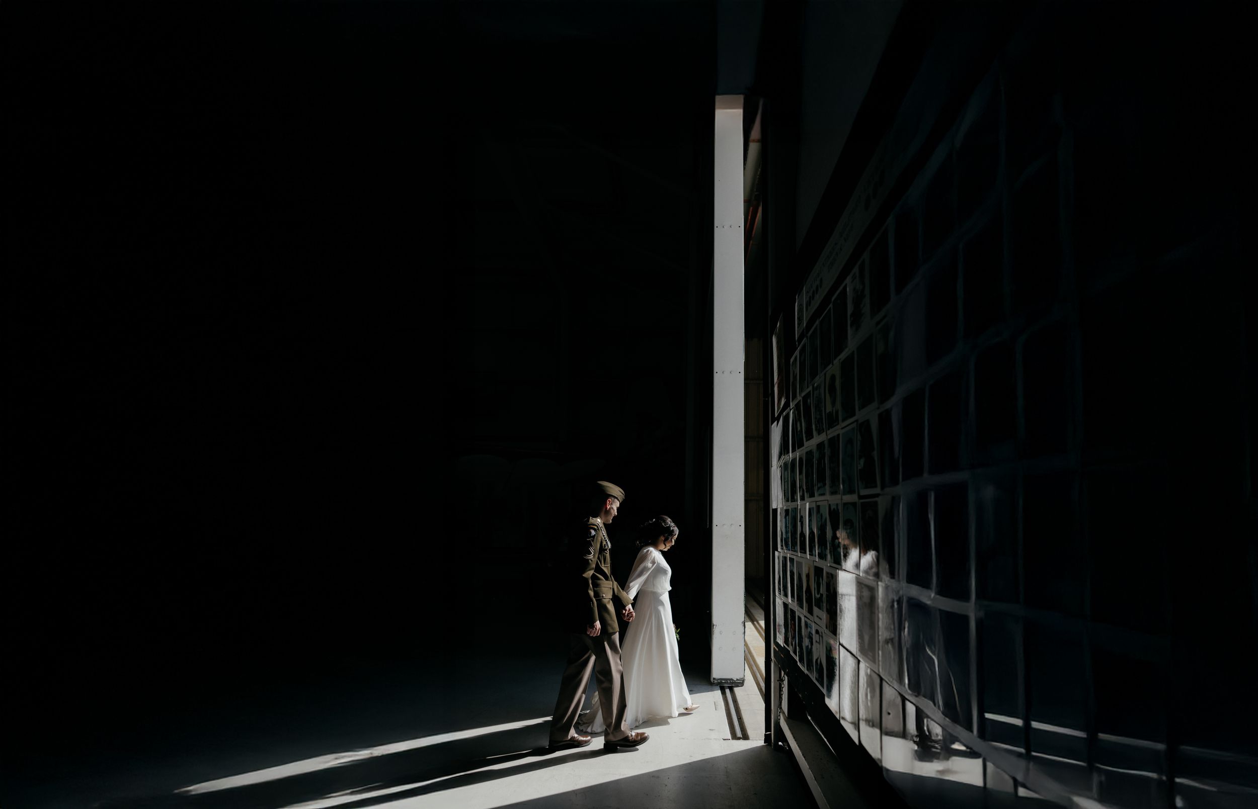 A couple in wedding attire walks through a dramatic beam of light against a dark architectural backdrop.
