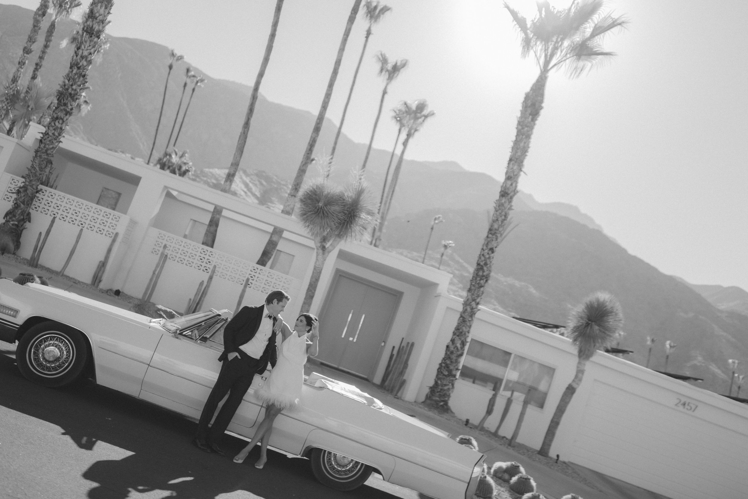 Black and white photo of a couple sharing a romantic moment beside a vintage convertible with palm trees and mountains.