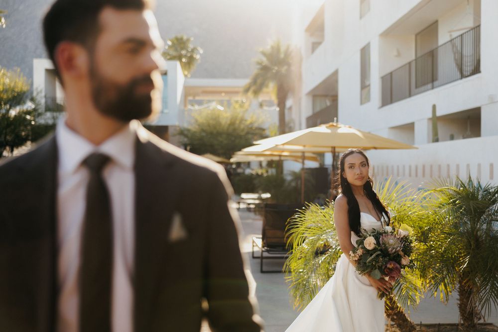 A couple in wedding attire stands in an outdoor venue with palm trees and white buildings in warm sunlight.