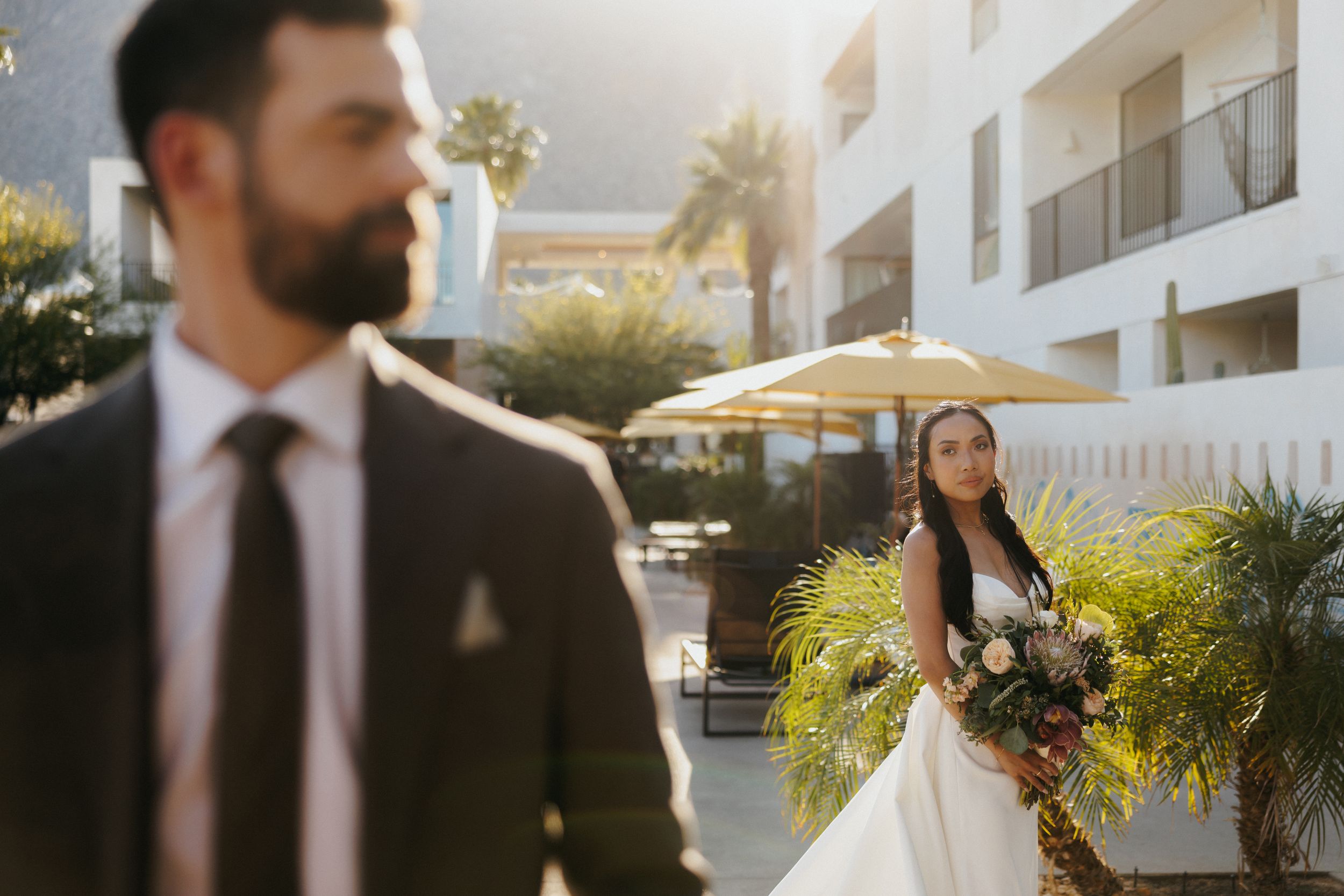 A couple in wedding attire stands in an outdoor venue with palm trees and white buildings in warm sunlight.
