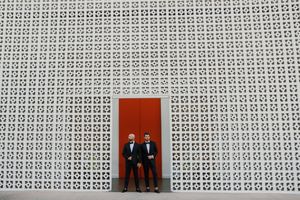 Two grooms in tuxedos stand before a geometric white wall with a bold red door at their wedding.