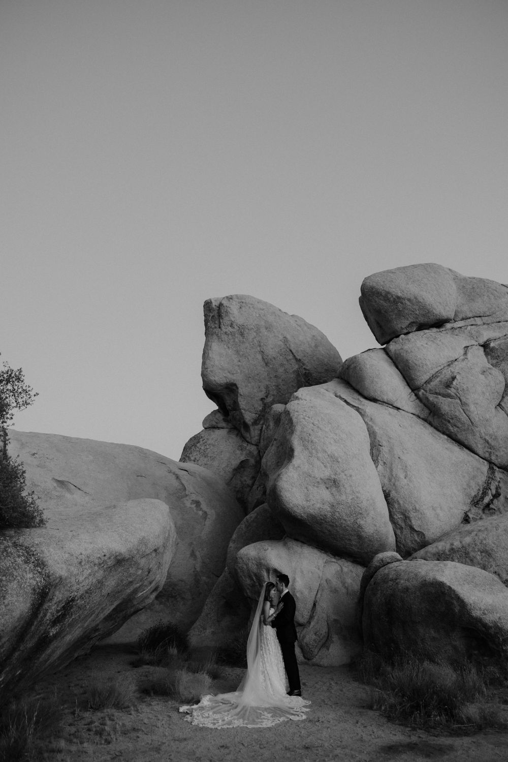A black and white wedding photo of a couple standing between large desert rock formations at Joshua Tree National Park.