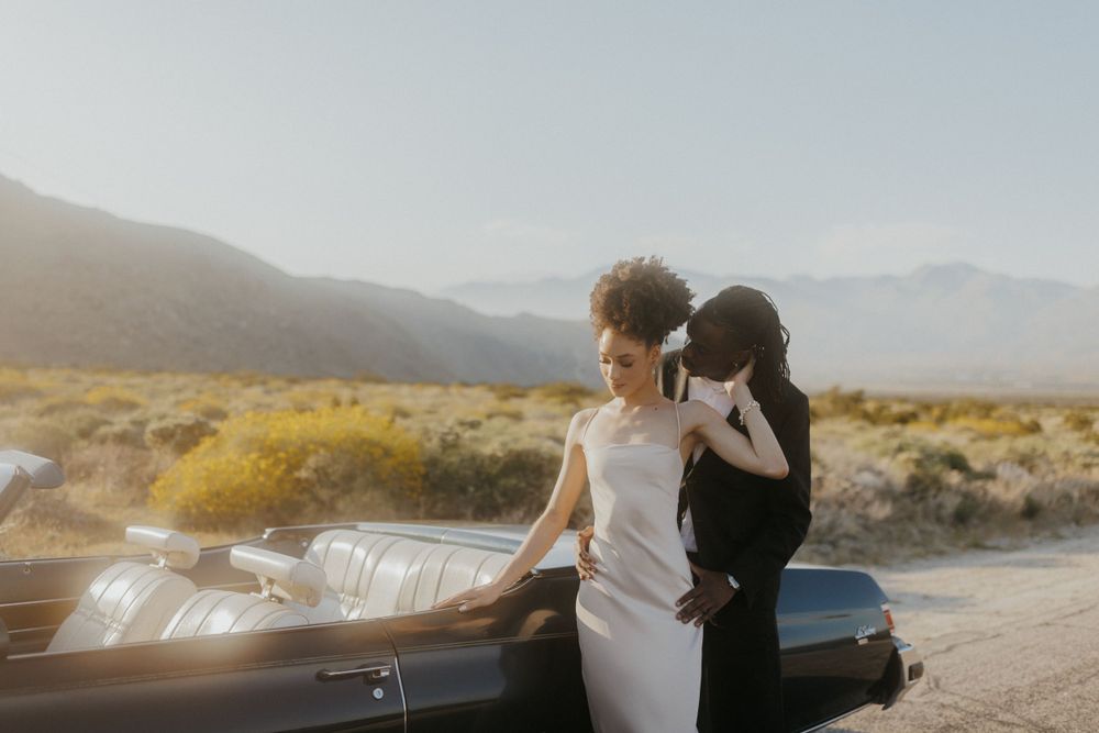 A vintage convertible car parked on a desert road at sunset with mountains in the background.