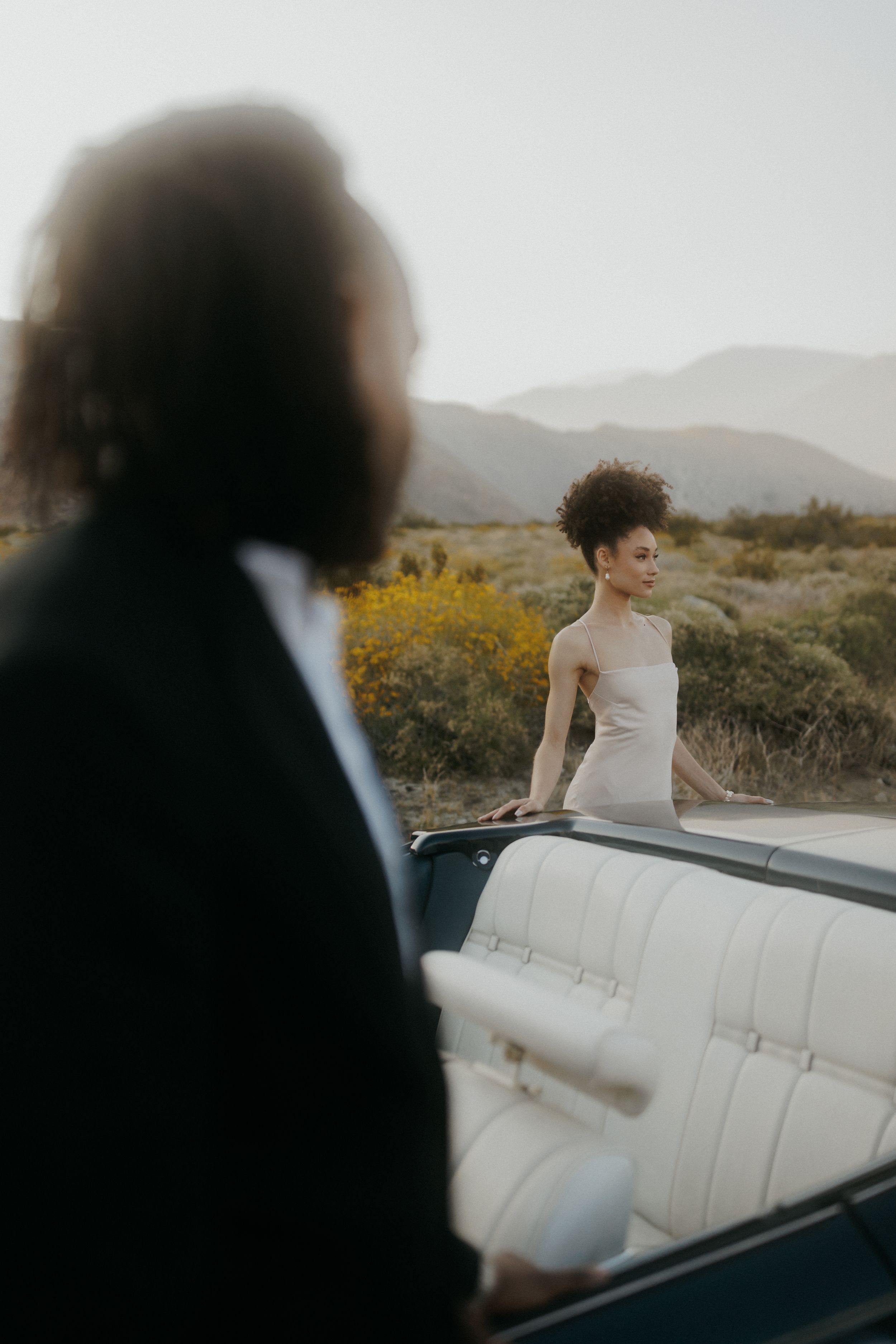 A couple in wedding attire pose with their vintage convertible in a desert landscape at golden hour.