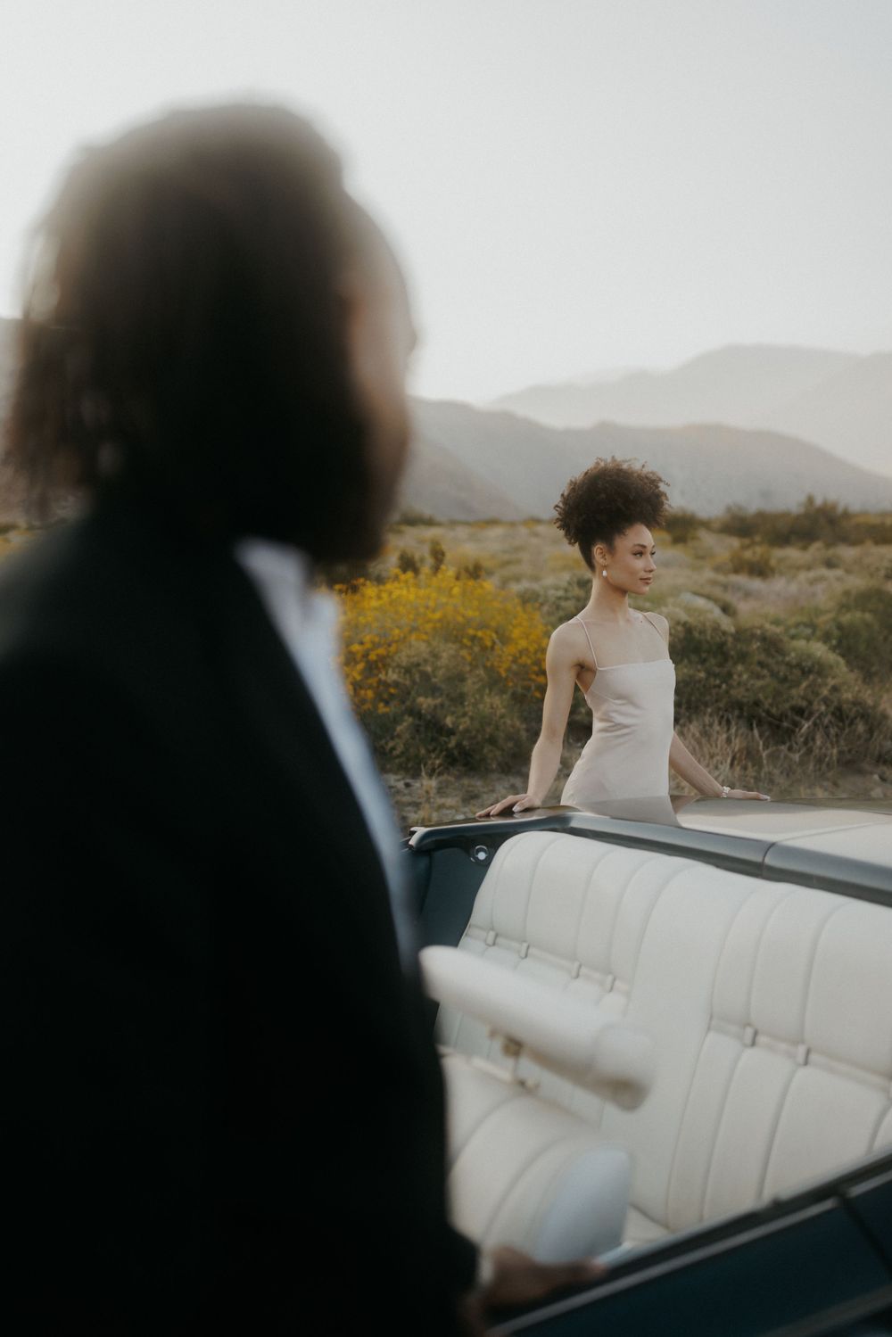 A couple in wedding attire pose with their vintage convertible in a desert landscape at golden hour.