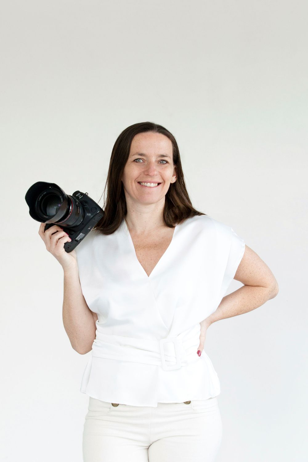 Photographe professionnel en tenue blanche tenant un appareil photo reflex numérique sur fond clair.
