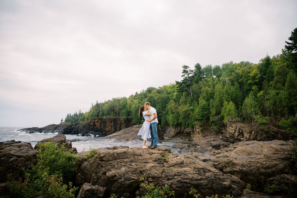 A couple embraces on rugged coastal cliffs overlooking Lake Superior with dense forest in the background.