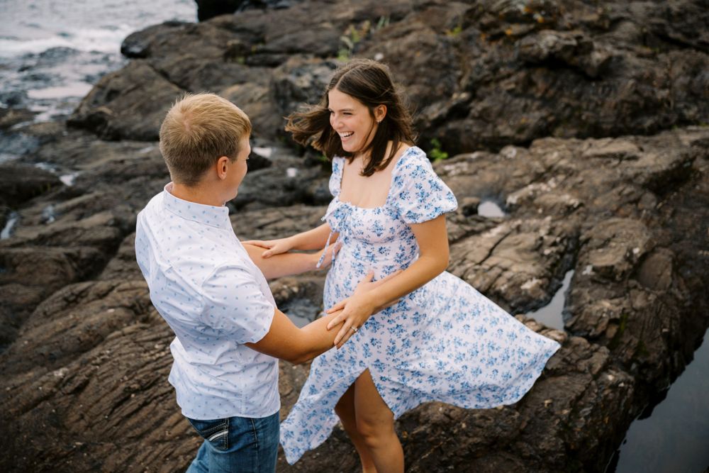 A couple in light blue and white outfits share a romantic moment on rocky coastal cliffs by the ocean.
