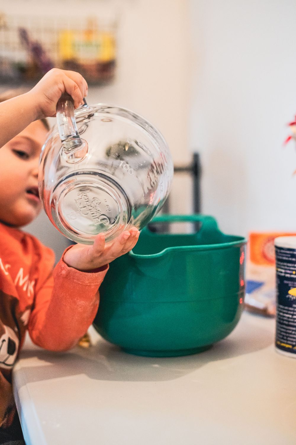 Small child carefully pouring liquid from glass pitcher into green bowl.