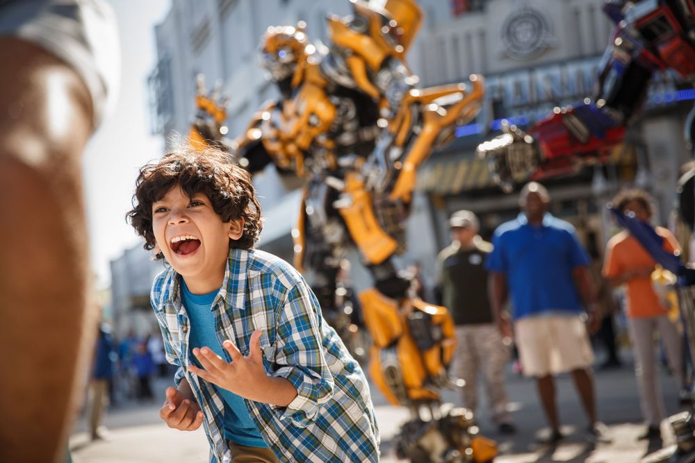 A young boy shows excitement while visiting an amusement park with large robot characters in the background.