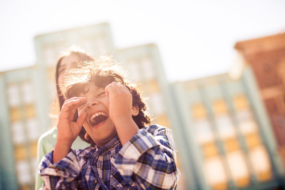 Child in plaid shirt laughs joyfully while playing outdoors on a sunny day with buildings in background.