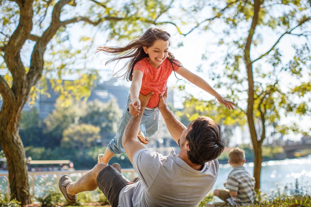 Parent and child enjoy playful outdoor moment in sunny park setting near water.
