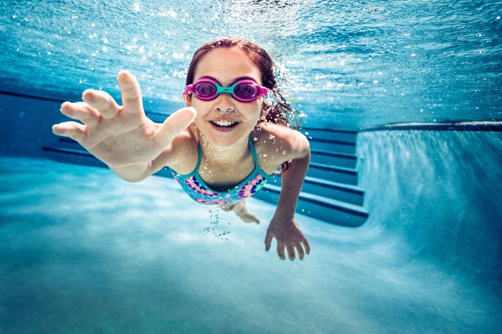Young swimmer with pink goggles reaches toward camera underwater in bright blue swimming pool.
