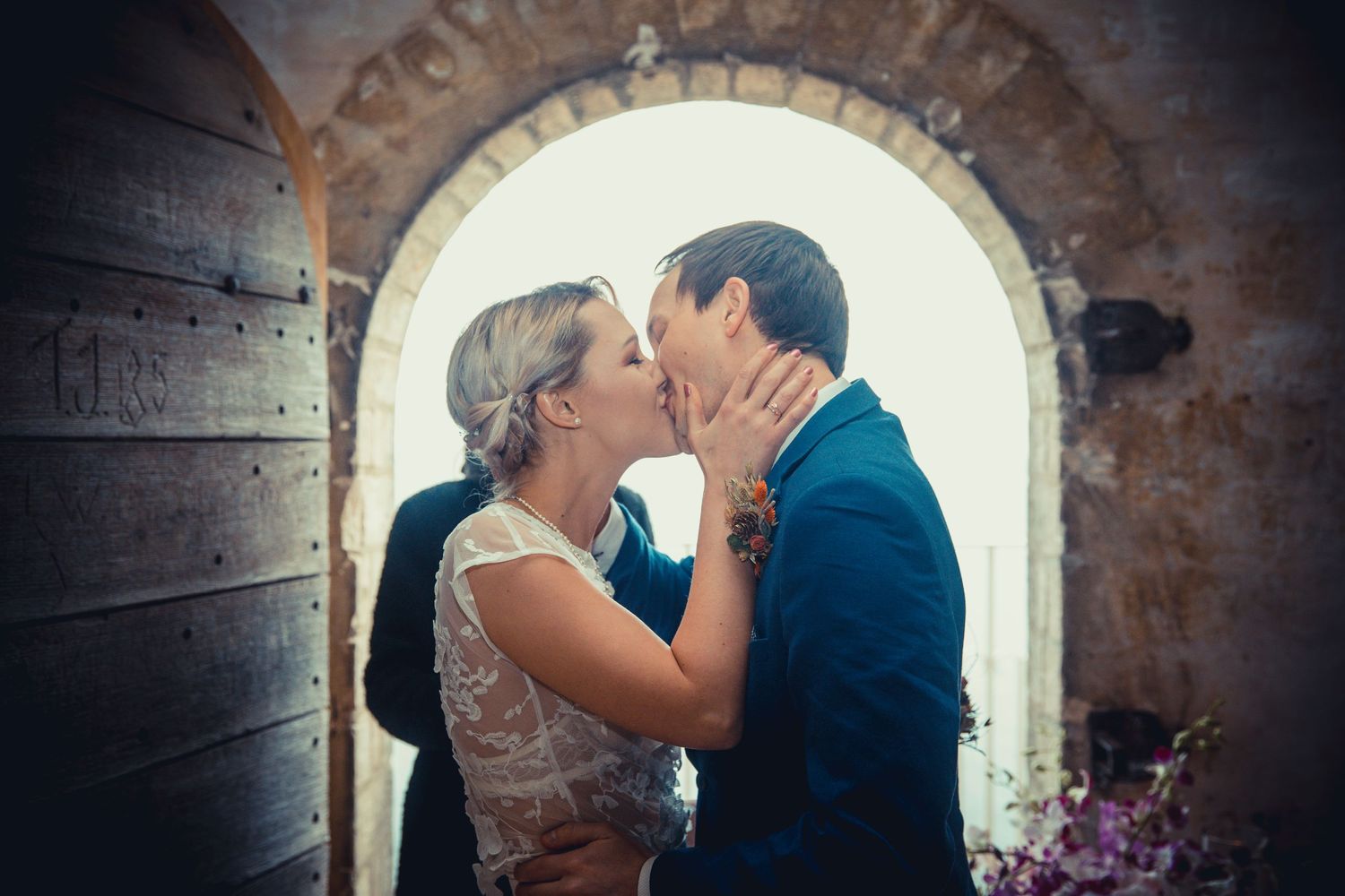 Intimate wedding kiss framed by stone archway with bright natural lighting.