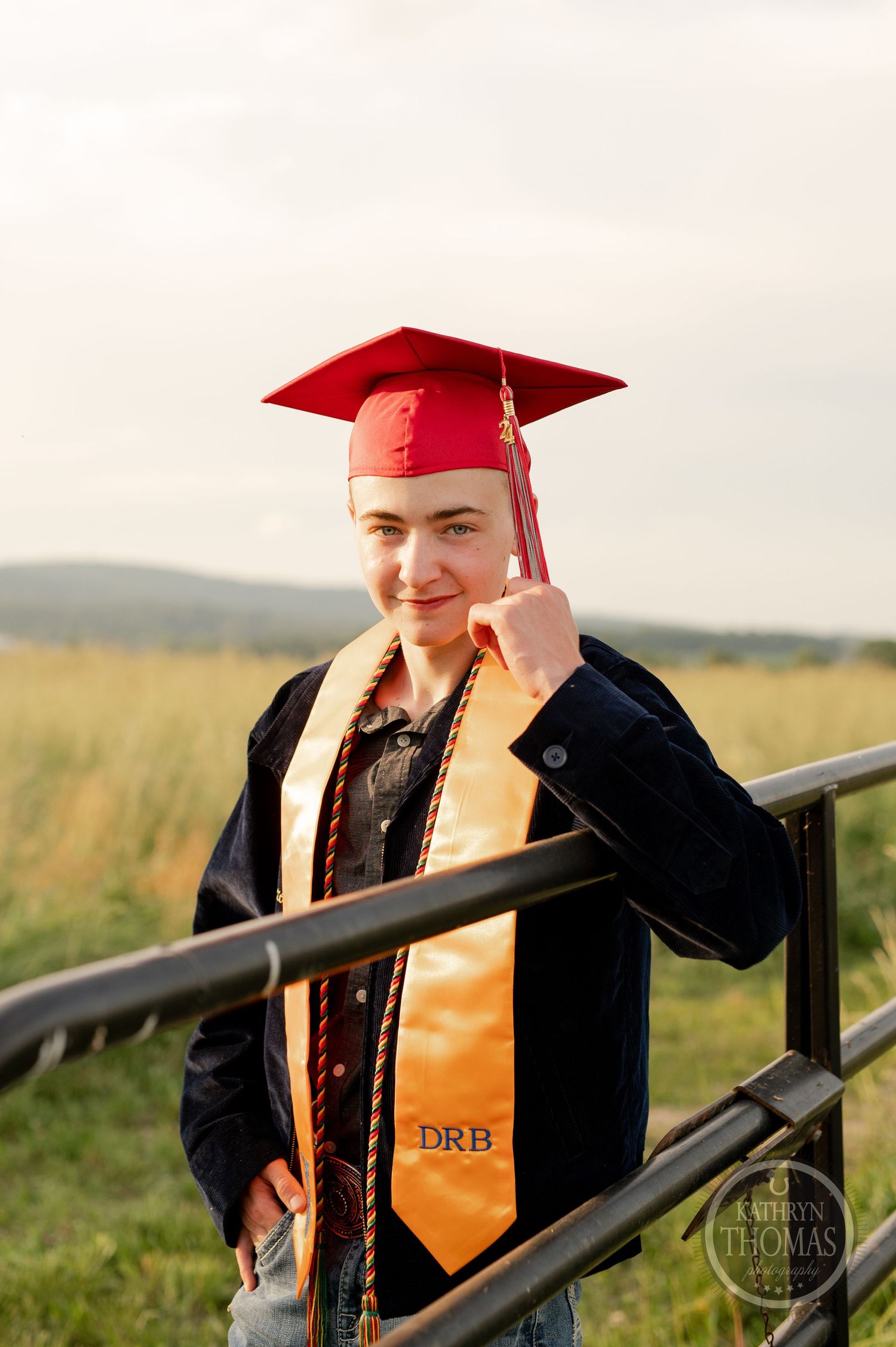 Graduate in red cap and gown with orange sash stands near railing in outdoor rural setting.