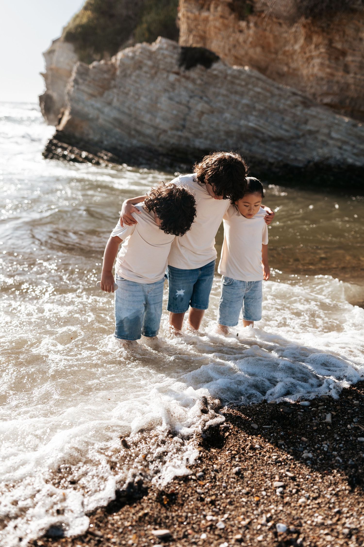 three young children standing on a beach while waves crash at their feet and they are looking down at the waves