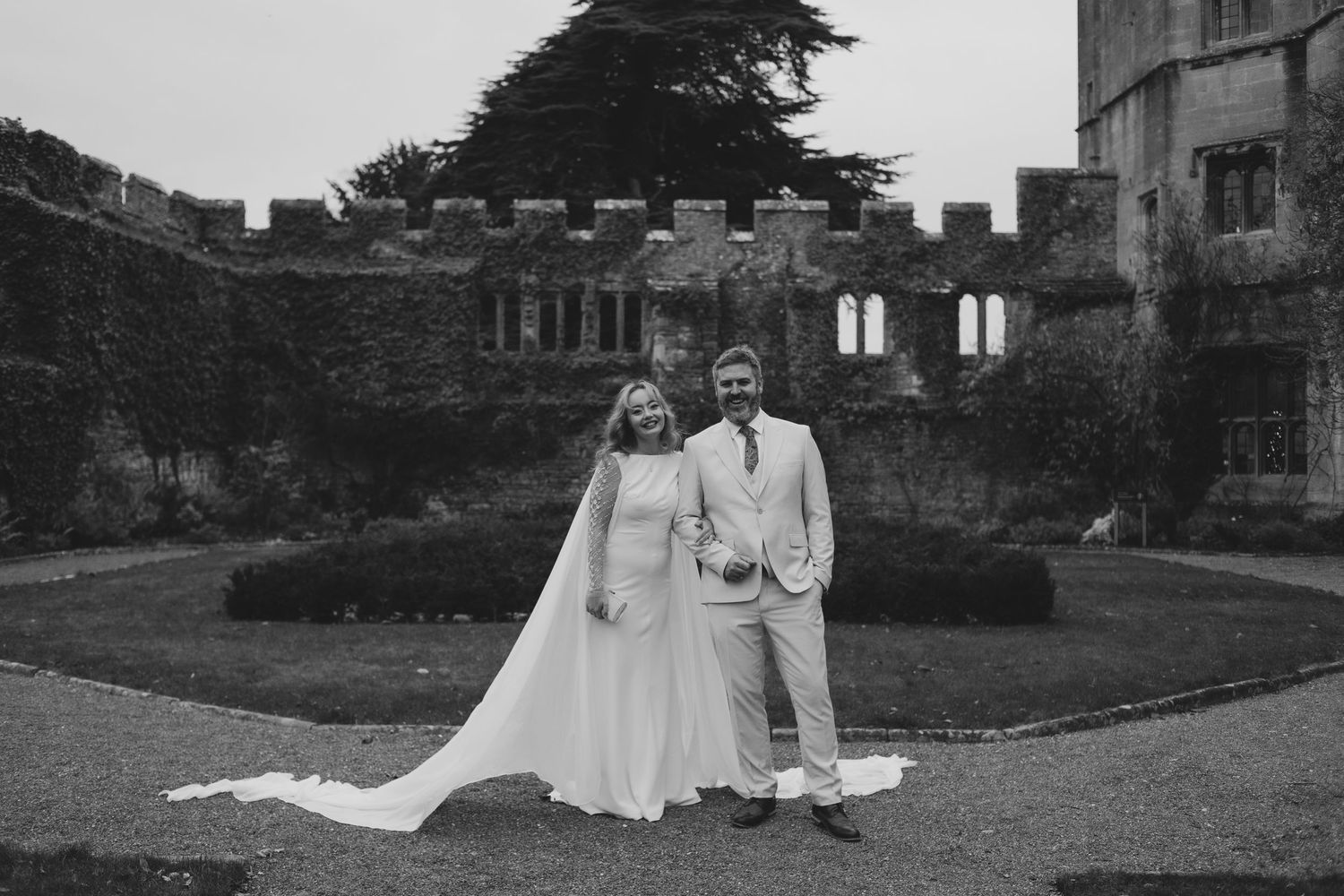 Candid black and white shot of newlyweds playfully posing with wedding dress on a garden lawn.