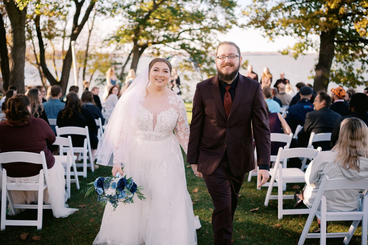 A bride and groom walk joyfully down the outdoor wedding aisle as guests look on from white folding chairs.