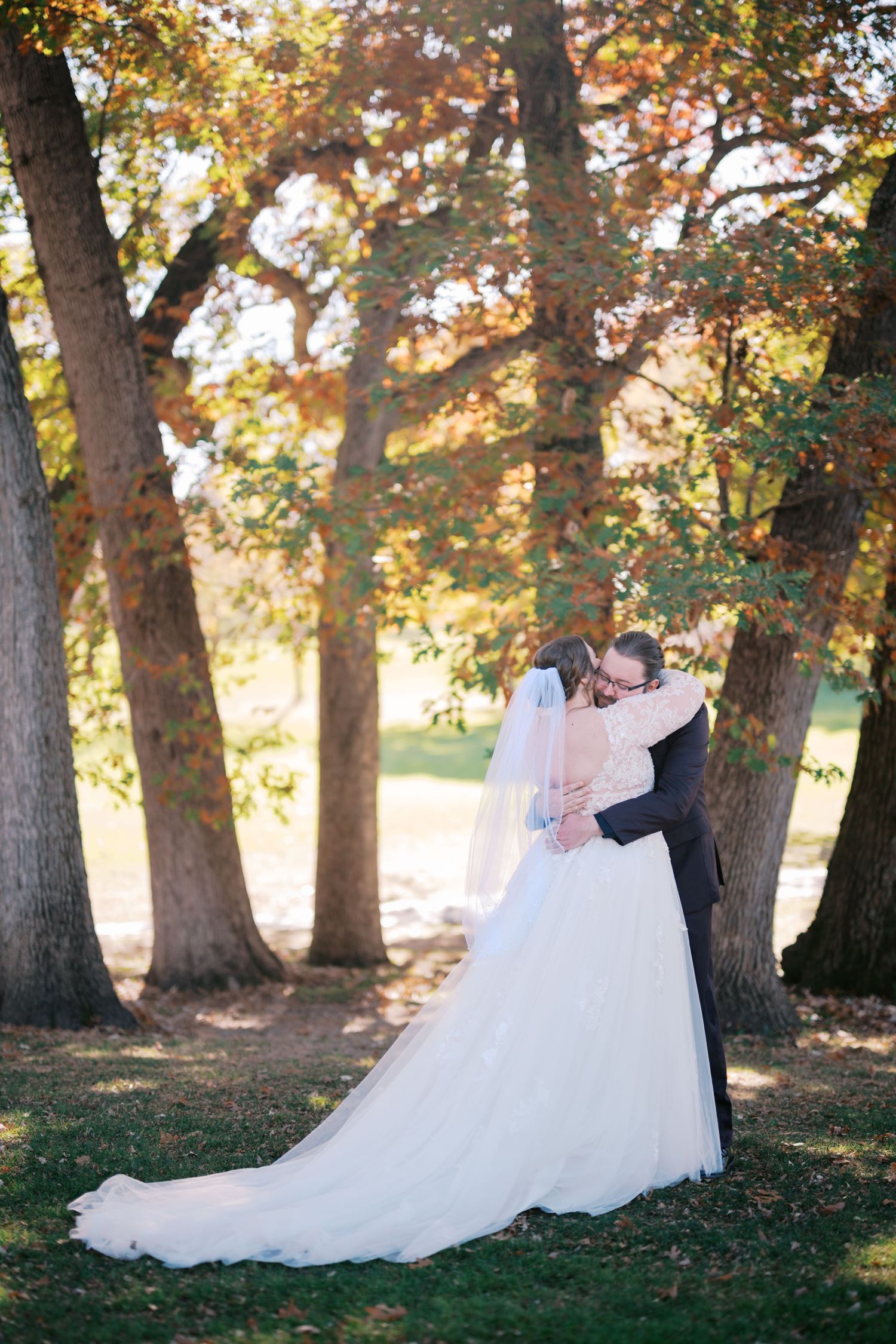 A couple shares an intimate moment under autumn trees in a wedding photo, her flowing white dress trailing behind them.