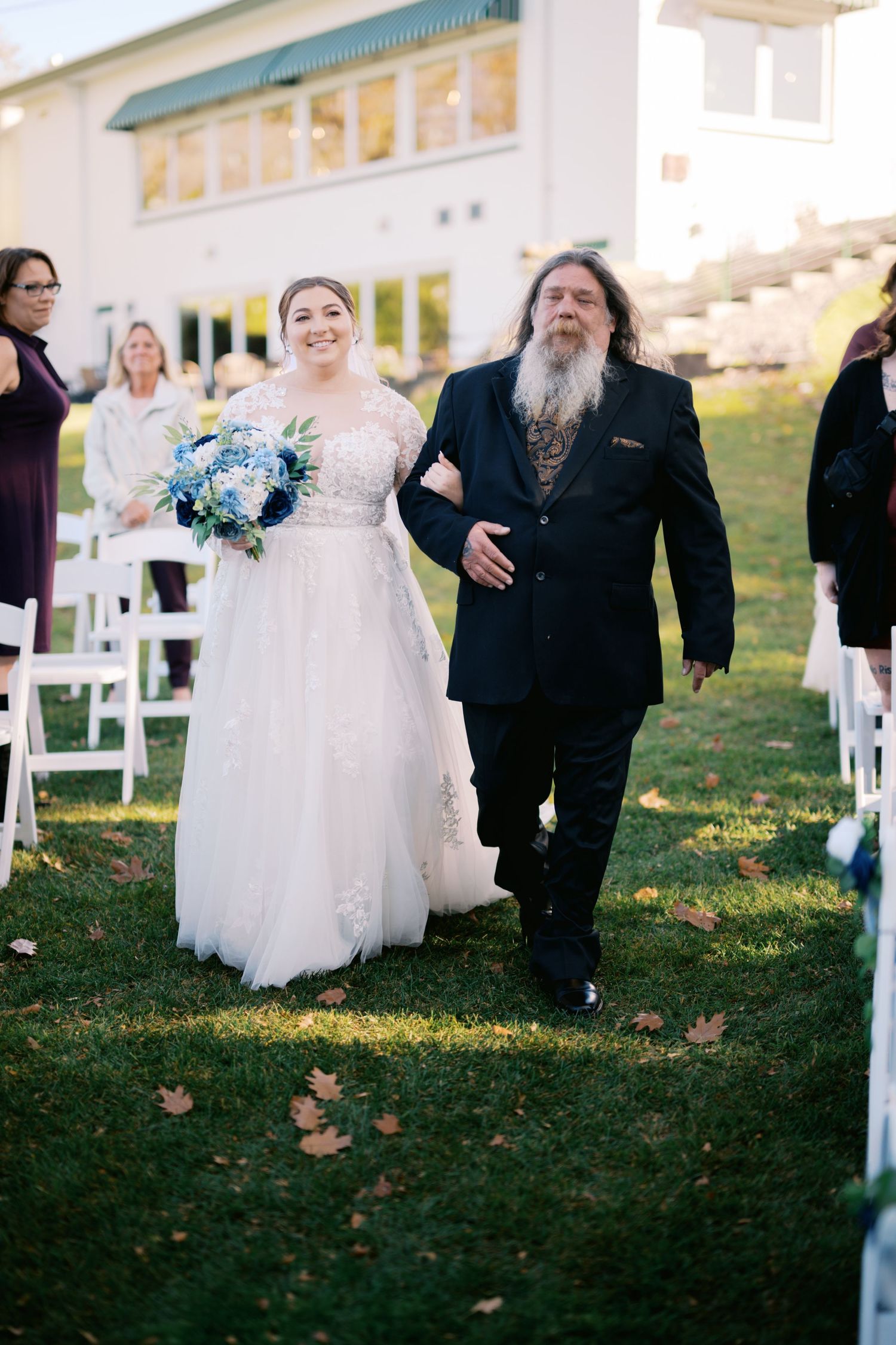 A bride in a white gown walks down the aisle with a man in a black suit at an outdoor wedding ceremony.